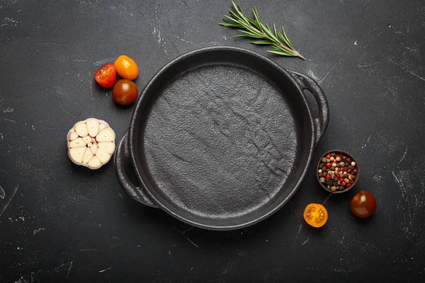 Taze sarımsak, domates, biberiye ve karışık biberiye ile çevrili koyu bir zemin üzerine demir tava dökmek