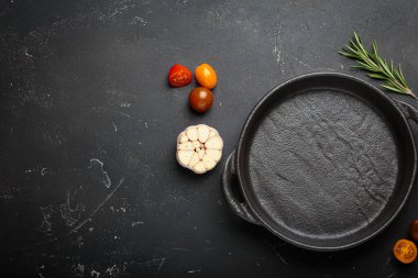 Taze sarımsak, domates, biberiye ve karışık biberiye ile çevrili koyu bir zemin üzerine demir tava dökmek