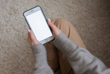 Genç bir kız beyaz bir halıda otururken elinde beyaz bir ekranla telefonu tutuyor. Yukarıdan görünüm.