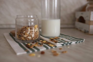 Müsli şeffaf bir kavanozda, Kunjon masasındaki bir masanın üzerinde duran bir şişe sütün yanında duruyor. Bir şişe süt, kavanozda mısır gevreği, masaya serpiştirilmiş tahıllar. Seçili odak.