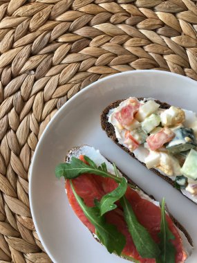 Hasır bir standın üzerinde beyaz bir tabak sandviç duruyor. Kırmızı balıklı sandviç, domates, roka, salata. seçili odak