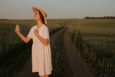Beyaz elbiseli ve şapkalı bir kız toprak bir yolda yeşil bir tarlada duruyor. Böceklerden, sivrisineklerden ve sineklerden püskürtülmüş bir sprey sıkarak..
