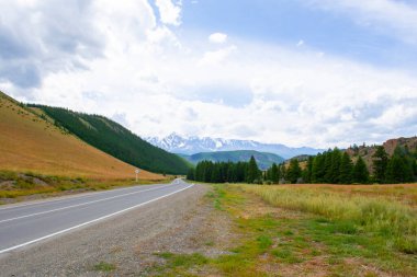 Dağların karlı zirveleri bulutların arka planında ve yol mesafeye kadar uzanıyor.