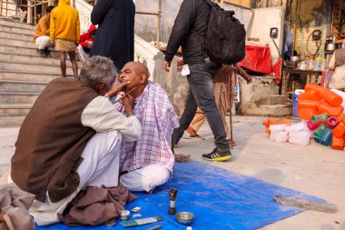 HARIDWAR, UTTARAKHAND, INDIA - Şubat 2021: Haridwar caddesinde kimliği belirsiz bir berber Har ki pauri ghat yakınındaki müşteriyi Kumbh Mela sırasında Hindu halkının ritüelinin bir parçası olarak tıraş ediyor.