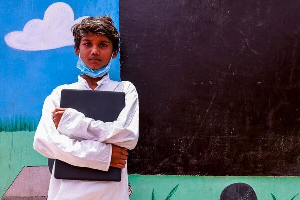 Portrait of a young Indian student from rural area standing with holding laptop in hands and medical mask on face near blackboard in the open school for education.