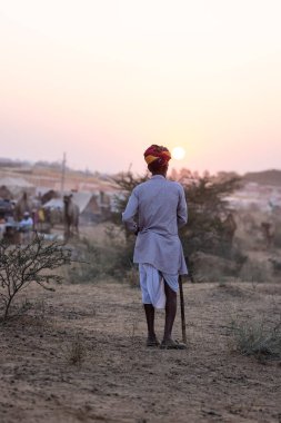 Puşkar, Rajasthan, Hindistan - 10 Kasım 2024: Geleneksel beyaz giysili rajasthani erkek portresi çöl panayırında renkli türbanla deve pazarında develeri takas ederken.