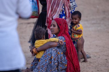 Puşkar, Rajasthan, Hindistan - 10 Kasım 2024: Kimliği belirsiz Hintli bir Rajasthani kadının portresi Renkli geleneksel elbiseli, çocuklarla birlikte kış sabahı Pushkar Deve Fuarı 'nda oturuyor.