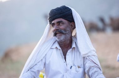 Puşkar, Rajasthan, Hindistan - 11 Kasım 2024: Geleneksel beyaz giysili rajasthani erkek portresi çöl panayırında renkli türbanla deve pazarında deve ticareti yaparken.