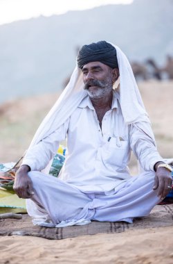 Puşkar, Rajasthan, Hindistan - 11 Kasım 2024: Geleneksel beyaz giysili rajasthani erkek portresi çöl panayırında renkli türbanla deve pazarında deve ticareti yaparken.