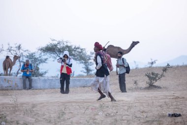Puşkar, Rajasthan, Hindistan - 11 Kasım 2024: Geleneksel beyaz giysili rajasthani erkek portresi çöl panayırında renkli türbanla deve pazarında deve ticareti yaparken.