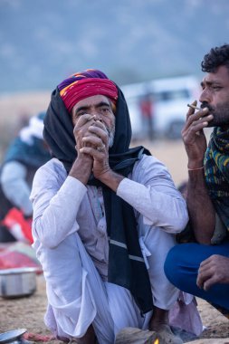 Puşkar, Rajasthan, Hindistan - 11 Kasım 2024: Geleneksel beyaz giysili rajasthani erkek portresi çöl panayırında renkli türbanla deve pazarında deve ticareti yaparken.