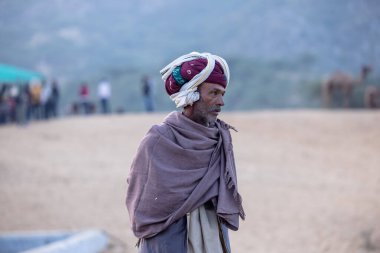 Puşkar, Rajasthan, Hindistan - 11 Kasım 2024: Geleneksel beyaz giysili rajasthani erkek portresi çöl panayırında renkli türbanla deve pazarında deve ticareti yaparken.