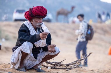 Puşkar, Rajasthan, Hindistan - 11 Kasım 2024: Geleneksel beyaz giysili rajasthani erkek portresi çöl panayırında renkli türbanla deve pazarında deve ticareti yaparken.