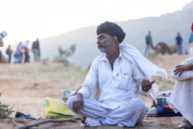 Puşkar, Rajasthan, Hindistan - 11 Kasım 2024: Geleneksel beyaz giysili rajasthani erkek portresi çöl panayırında renkli türbanla deve pazarında deve ticareti yaparken.