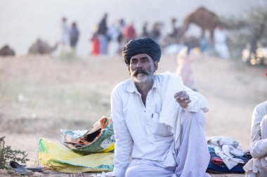 Puşkar, Rajasthan, Hindistan - 11 Kasım 2024: Geleneksel beyaz giysili rajasthani erkek portresi çöl panayırında renkli türbanla deve pazarında deve ticareti yaparken.