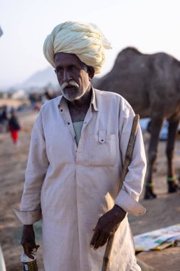 Puşkar, Rajasthan, Hindistan - 11 Kasım 2024: Geleneksel beyaz giysili rajasthani erkek portresi çöl panayırında renkli türbanla deve pazarında deve ticareti yaparken.