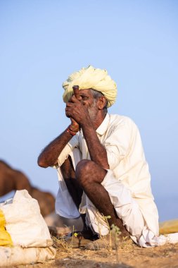 Puşkar, Rajasthan, Hindistan - 11 Kasım 2024: Geleneksel beyaz giysili rajasthani erkek portresi çöl panayırında renkli türbanla deve pazarında deve ticareti yaparken.