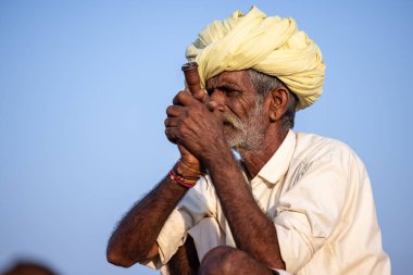 Puşkar, Rajasthan, Hindistan - 11 Kasım 2024: Geleneksel beyaz giysili rajasthani erkek portresi çöl panayırında renkli türbanla deve pazarında deve ticareti yaparken.