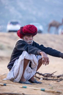 Puşkar, Rajasthan, Hindistan - 11 Kasım 2024: Geleneksel beyaz giysili rajasthani erkek portresi çöl panayırında renkli türbanla deve pazarında deve ticareti yaparken.