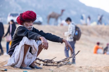 Puşkar, Rajasthan, Hindistan - 11 Kasım 2024: Geleneksel beyaz giysili rajasthani erkek portresi çöl panayırında renkli türbanla deve pazarında deve ticareti yaparken.