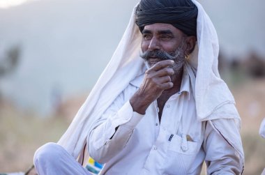 Puşkar, Rajasthan, Hindistan - 11 Kasım 2024: Geleneksel beyaz giysili rajasthani erkek portresi çöl panayırında renkli türbanla deve pazarında deve ticareti yaparken.