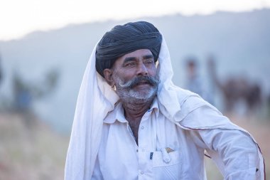 Puşkar, Rajasthan, Hindistan - 11 Kasım 2024: Geleneksel beyaz giysili rajasthani erkek portresi çöl panayırında renkli türbanla deve pazarında deve ticareti yaparken.