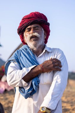 Puşkar, Rajasthan, Hindistan - 11 Kasım 2024: Geleneksel beyaz giysili rajasthani erkek portresi çöl panayırında renkli türbanla deve pazarında deve ticareti yaparken.