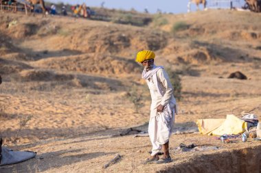 Puşkar, Rajasthan, Hindistan - 11 Kasım 2024: Geleneksel beyaz giysili rajasthani erkek portresi çöl panayırında renkli türbanla deve pazarında deve ticareti yaparken.