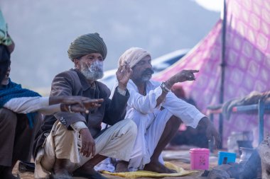 Puşkar, Rajasthan, Hindistan - 11 Kasım 2024: Geleneksel beyaz giysili rajasthani erkek portresi çöl panayırında renkli türbanla deve pazarında deve ticareti yaparken.