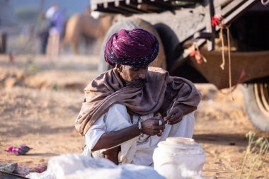 Puşkar, Rajasthan, Hindistan - 11 Kasım 2024: Geleneksel beyaz giysili rajasthani erkek portresi çöl panayırında renkli türbanla deve pazarında deve ticareti yaparken.