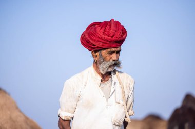 Puşkar, Rajasthan, Hindistan - 11 Kasım 2024: Çöldeki fuar alanında deve ticaret fuarı sırasında renkli türbanlı geleneksel beyaz giysili bir rajasthani erkeğinin portresi.