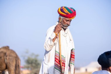 Puşkar, Rajasthan, Hindistan - 11 Kasım 2024: Çöldeki fuar alanında deve ticaret fuarı sırasında renkli türbanlı geleneksel beyaz giysili bir rajasthani erkeğinin portresi.