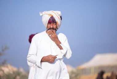 Puşkar, Rajasthan, Hindistan - 11 Kasım 2024: Çöldeki fuar alanında deve ticaret fuarı sırasında renkli türbanlı geleneksel beyaz giysili bir rajasthani erkeğinin portresi.