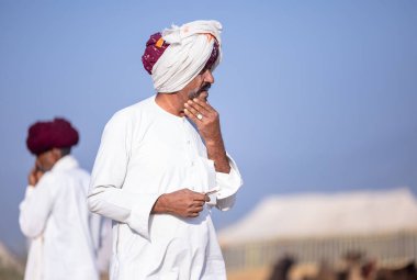 Puşkar, Rajasthan, Hindistan - 11 Kasım 2024: Çöldeki fuar alanında deve ticaret fuarı sırasında renkli türbanlı geleneksel beyaz giysili bir rajasthani erkeğinin portresi.