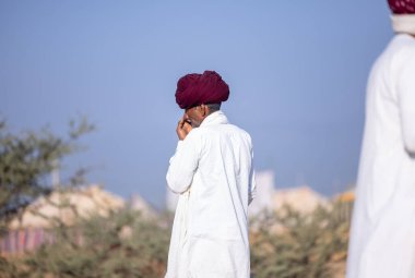 Puşkar, Rajasthan, Hindistan - 11 Kasım 2024: Çöldeki fuar alanında deve ticaret fuarı sırasında renkli türbanlı geleneksel beyaz giysili bir rajasthani erkeğinin portresi.