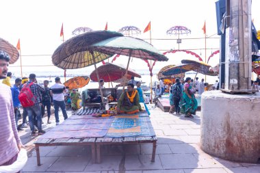 Varanasi, Uttar Pradesh, Hindistan - 22 Mart 2024: Varanasi 'deki çetelerin yakınındaki assi ghat' ta şemsiye altında oturan geleneksel bir Brahman rahibinin portresi.