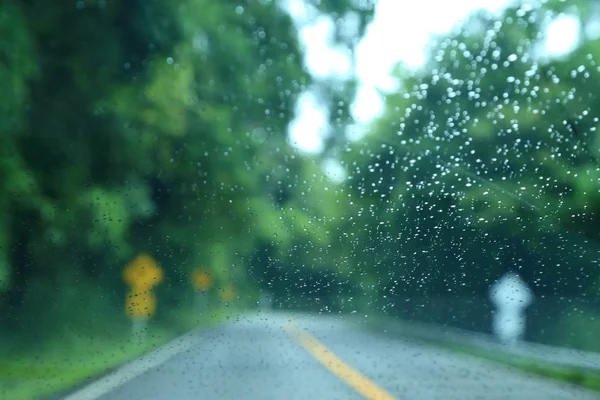 Blurred road in the forest look through rain drop on car glass. — Stock Photo, Image