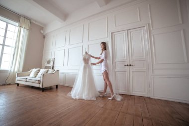 Gelin sabahı. Beyaz cüppeli ve peçeli bir kız gelinliğin yanında duruyor.