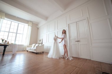 Gelin sabahı. Beyaz cüppeli ve peçeli bir kız beyaz bir gelinliğin yanında duruyor.