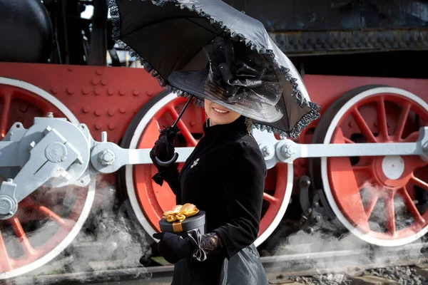 Beautiful girl in a black dress and hat near an old steam locomotive and big iron wheels. Blond beauty. Vintage portrait of the last century, retro journey. — Stock Photo, Image