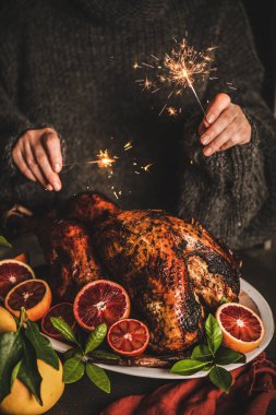 Yünlü kazaklı bir kadın Noel 'de ışıltı yakıyor ya da bayram sofrası için portakallı tavuk pişiriyor. Noel ve Yeni Yıl bayramı kutlamaları ve geleneksel yemekler