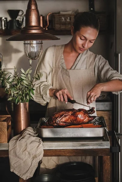 Keten önlüklü genç bir kadın Şükran Günü tatili için kızarmış ördek oyuyor ya da modern mutfak içinde Noel partisi kutlaması yapıyor. Geleneksel Sonbahar ya da Kış tatili konforu
