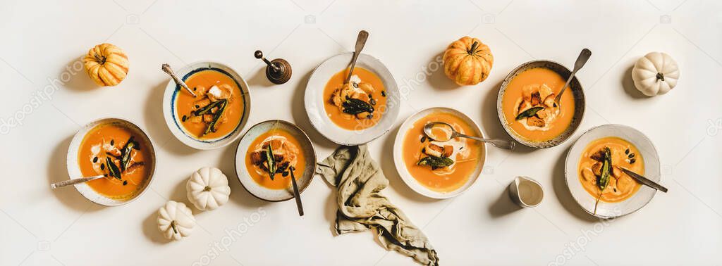 Flat-lay of homemade fall pumpkin cream soup in plates with sour cream, pumpkin seeds and bread croutons over plain white table with fresh pumpkins and spices, top view. Healthy comfort food