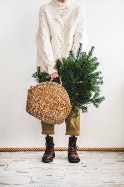 Beyaz yün süveter, haki rengi pantolon ve çizme giymiş insan figürü. Beyaz arka planda elde dekorasyon için her zaman yeşil Noel ağacı dallarıyla dolu hasır bir çanta tutuyor. Noel, Yeni Yıl
