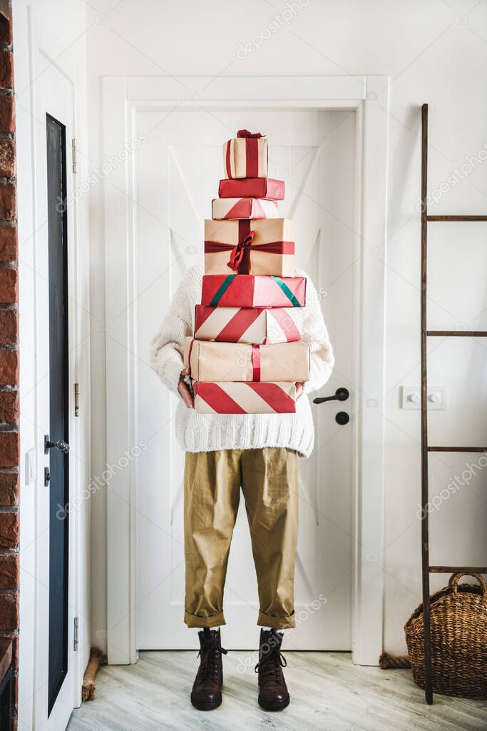Human figure in winter sweater, khaki pants and boots holding heap of Christmas festive gift boxes with red ribbon and bows, home interior at background. Christmas, Boxing day, New year mood concept