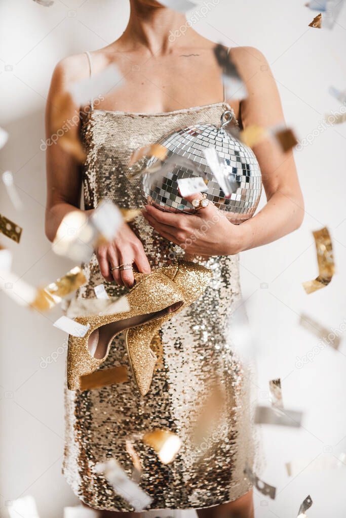 Young woman figure in festive glittering cocktail golden mini dress holding discoball and high heels shoes in hands over white wall with confetti. New Year holiday party concept