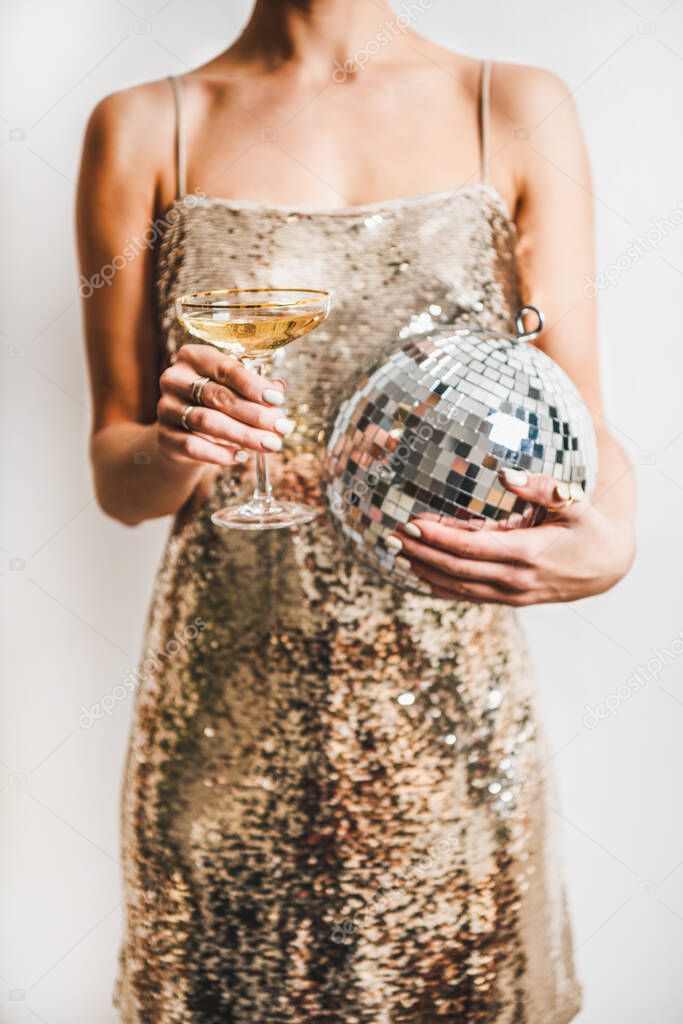 Young woman figure in festive glittering cocktail golden mini dress holding glass of champagne and discoball over white wall background. New Year holiday party concept