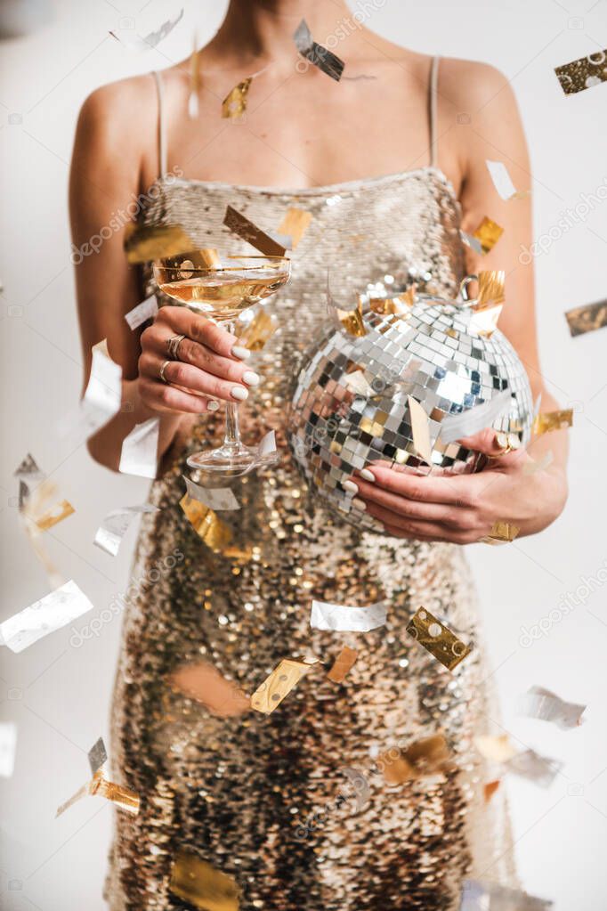 Young woman figure in festive glittering cocktail golden mini dress holding glass of champagne and discoball in cloud of golden confetti, white background. New Year holiday party concept