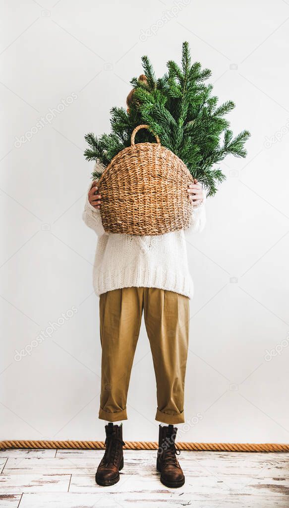 Female figure in white woolen winter sweater, khaki pants and boots standing holding wicker bag with evergreen Christmas tree branches for decoration at home over white background. Christmas, New year