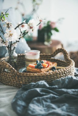 Yatakta kahvaltı. Kan portakalı ve yaban mersinli waffle, cam içinde gül latte ve sepet tepsisindeki beyaz çiçek, koyu gri keten çarşaflar, seçici odak, dikey kompozisyon
